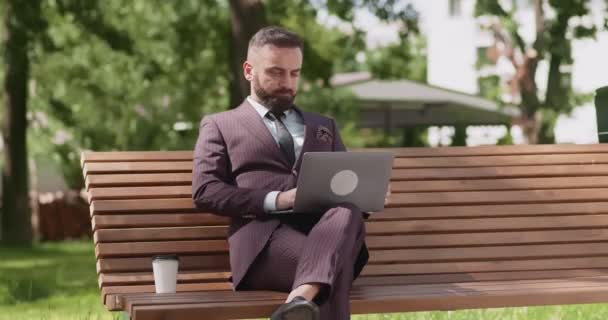 Wygodny harmonogram pracy. Dojrzały biznesmen pracujący na laptopie, siedzący na ławce w parku z kawą — Wideo stockowe