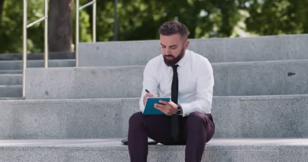 Бизнесмен средних лет сидит на лестнице в парке и пишет заметки, создавая новый бизнес-план по маркетинговой стратегии — стоковое видео