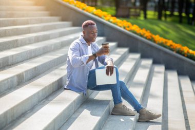 Tablet bilgisayarlı siyahi adam şehir merkezindeki taş merdivenlerde kahve içiyor.