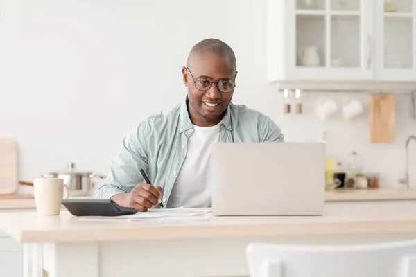 Σχέδιο οικονομικών. Θετικός αφροαμερικανός ώριμος άνθρωπος που υπολογίζει τους φόρους και χρησιμοποιεί φορητό υπολογιστή, κάθεται στην κουζίνα — Φωτογραφία Αρχείου