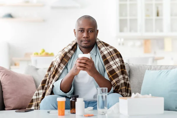 Homem negro coberto de cobertor sentindo-se mal, segurando caneca com bebida quente, sentado no sofá em casa interior, espaço de cópia — Fotografia de Stock