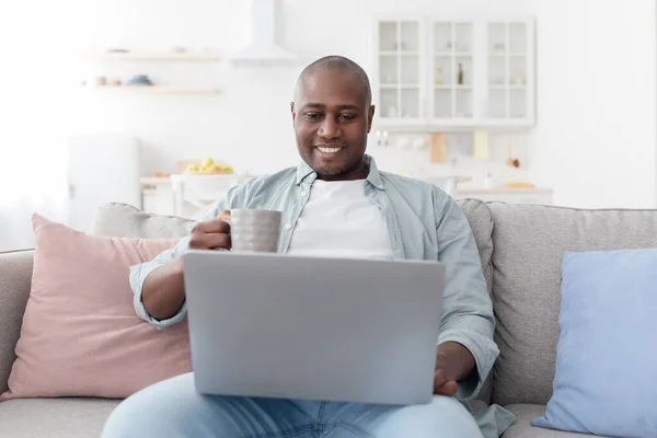 Leyendo noticias matutinas. Hombre afroamericano feliz usando el ordenador portátil y beber café caliente, descansando en el sofá — Foto de Stock