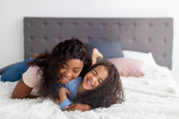 Kleines schwarzes Mädchen und ihre Mama umarmen sich zu Hause auf dem Bett, umarmen sich, lachen und haben Spaß zusammen — Stockfoto