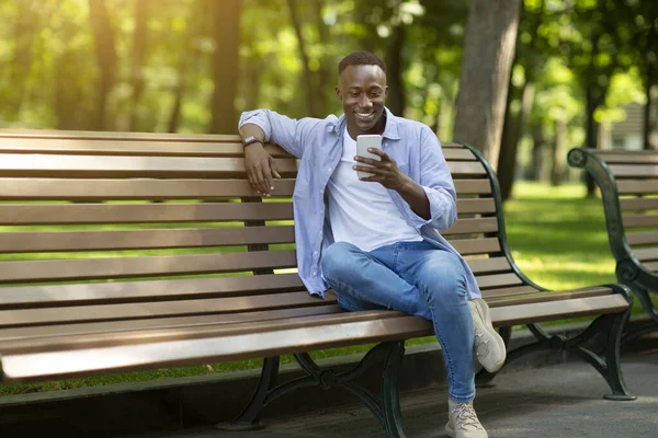 Sorrindo cara preto sentado no banco no parque e usando seu smartphone, espaço de cópia — Fotografia de Stock