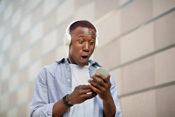 Шокований чорний чоловік в навушниках дивиться на екран мобільного телефону біля цегляної стіни на міській вулиці — стокове фото
