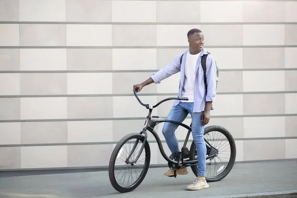 Aventura urbana. Homem preto feliz com mochila e bicicleta de pé perto da parede de tijolo ao ar livre e olhando para o lado, espaço cópia — Fotografia de Stock
