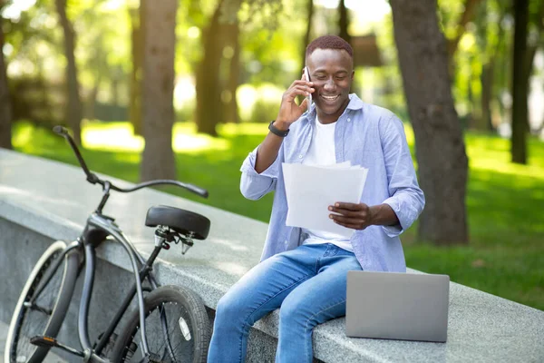 Homem afro-americano alegre com bicicleta e documentos discutindo questões de negócios no celular no parque — Fotografia de Stock