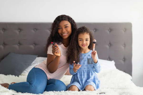 Siyahi anne ve güzel kızının makyaj ürünlerini tutarken ve yatakta kameraya gülümserken tam boy portresi. — Stok fotoğraf