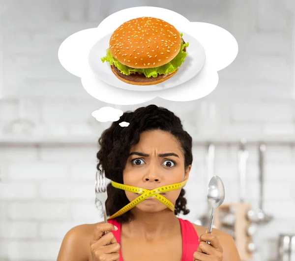 Concepto de pérdida de peso. Mujer afroamericana con cinta métrica alrededor de su boca soñando con hamburguesa malsana — Foto de Stock