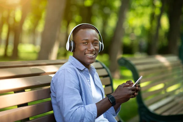 Красивий чорний чоловік розслабляється на лавці в парку і насолоджується своєю улюбленою музикою на смартфоні — стокове фото