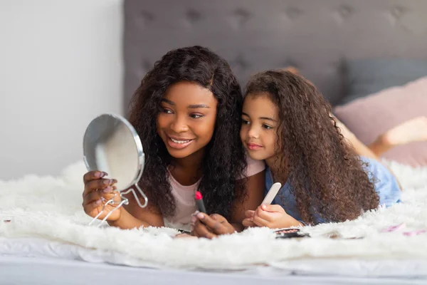 Χαρούμενη μαύρη γυναίκα και το μικρό της παιδί με προϊόντα μακιγιάζ κοιτάζοντας στον καθρέφτη στο κρεβάτι στο σπίτι — Φωτογραφία Αρχείου