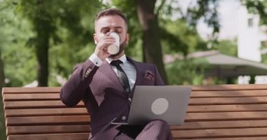 Kendine güvenen yetişkin iş adamı dizüstü bilgisayarla çalışıyor, bankta oturuyor ve kahve içiyor.