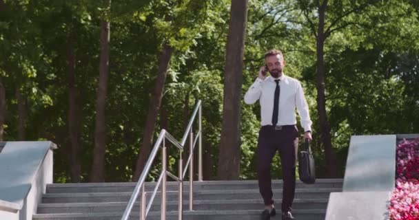 Namierzanie zajętego biznesmena chodzącego z torebką i rozmawiającego przez komórkę, idącego do biura przez park miejski — Wideo stockowe