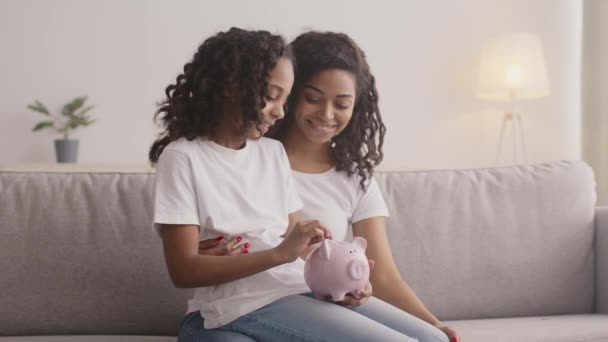 Finansiell kompetens för juniorer. Svart tjej sätter mynt i spargris, sitter på sina mödrars knän hemma — Stockvideo
