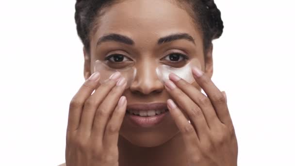 Neşeli, Afro-Amerikalı bir bayan gözlerinin altına altın rengi anti-yorgunluk bantları yapıştırıyor, kameraya gülümsüyor, beyaz arka plan — Stok video