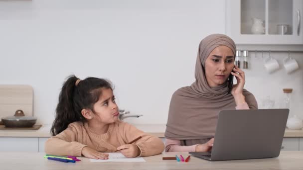 Madre musulmana trabajando en línea ignorando a su hija hablando por teléfono en interiores — Vídeos de Stock