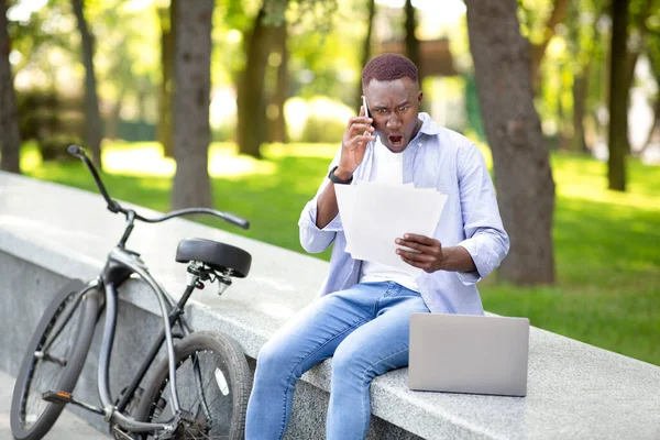 Εξοργισμένος Αφροαμερικανός με χαρτιά και λάπτοπ να ουρλιάζει στο κινητό του κοντά στο ποδήλατό του στο πάρκο. — Φωτογραφία Αρχείου