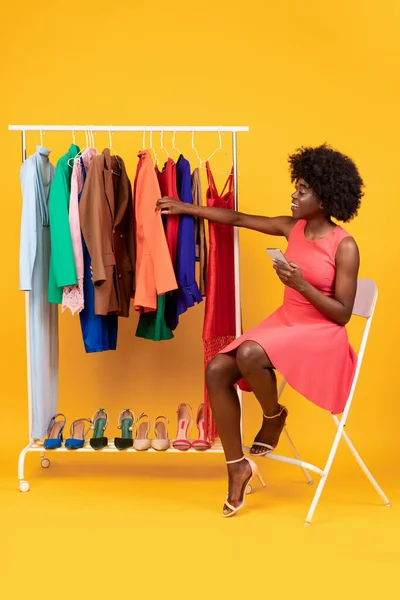 Feminino usando Smartphone escolhendo roupas na cremalheira de roupas, fundo amarelo — Fotografia de Stock