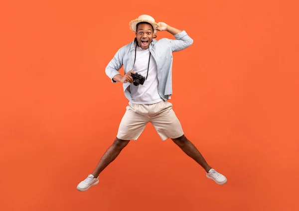 Teljes hossza izgatott fekete férfi nyári ruha tartó professzionális kamera és ugrás narancssárga stúdió háttér — Stock Fotó