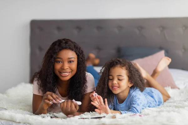 Fröhliche schwarze Mutter und ihre kleine Tochter bei Maniküre, zu Hause im Bett liegend — Stockfoto