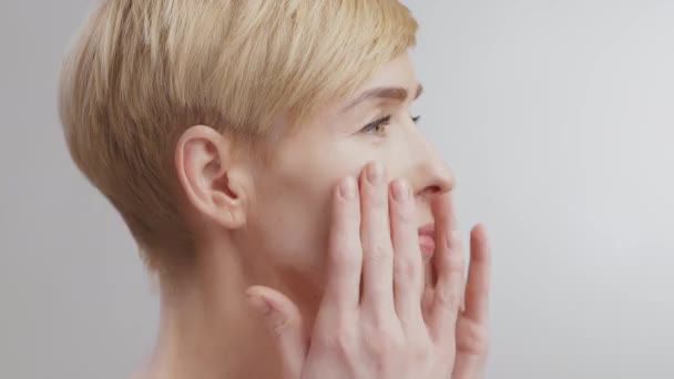 Profilporträtt av medelålders kvinna som masserar ansiktet, applicerar bortskämd lotion, vänder ansiktet mot kameran och ler — Stockvideo