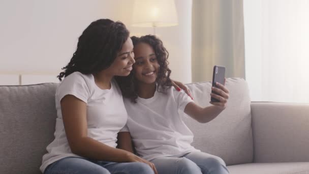 Selfie de família. Menina afro-americana bonito fazendo fotografia no telefone celular com a mãe, sentado no sofá em casa — Vídeo de Stock