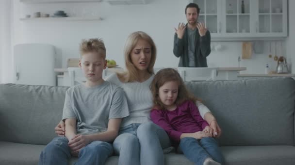 Nieszczęśliwa matka siedzi na kanapie obejmując smutne dzieci i płacze, zły człowiek krzyczy i kłóci się na tle — Wideo stockowe