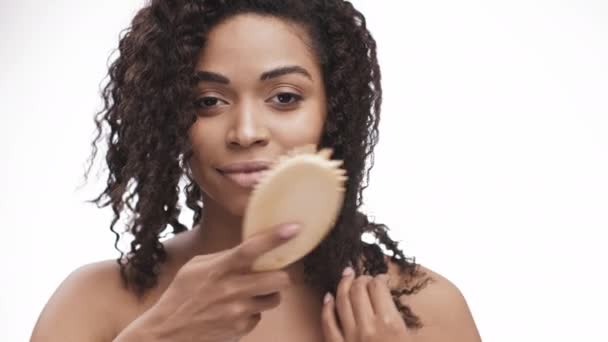 Feliz hermosa mujer afroamericana peinándose el pelo con cepillo de pelo, sonriendo a la cámara, fondo de estudio blanco — Vídeo de stock