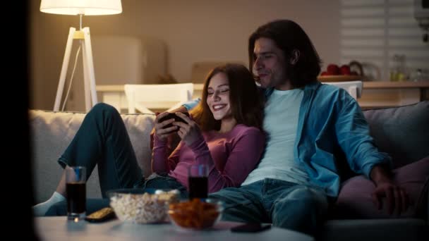若いです幸せな男と女は自宅でソファに休んで、スマホでビデオを見ますスナックと一緒に — ストック動画
