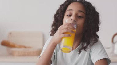 Tatlı Afro-Amerikalı genç kız taze portakal suyunu bardaktan içiyor ve kameraya gülümsüyor, mutfakta oturuyor.