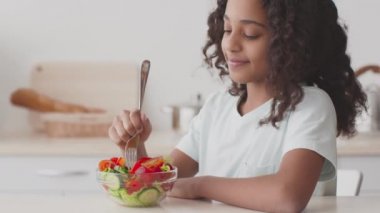 Sağlıklı bir akşam yemeği. Şirin Afrikalı Amerikalı genç kız taze sebze salatasının tadını çıkarıyor, mutfakta oturuyor, fotoğraf çekiyor.