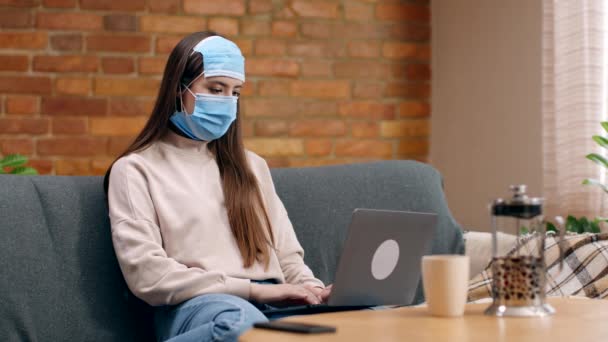 Fiatal nő visel három védő maszk az arcon gazdaság laptop, felkészülés a videohívás, ül a kanapén otthon — Stock videók