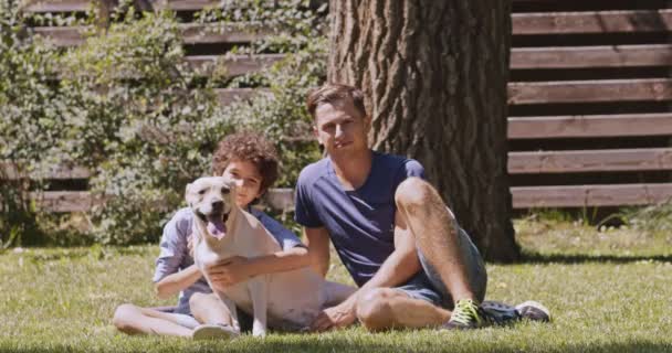 快乐的家庭年轻的男子和少年与狗坐在户外，对着相机微笑，躺在绿草上 — 图库视频影像