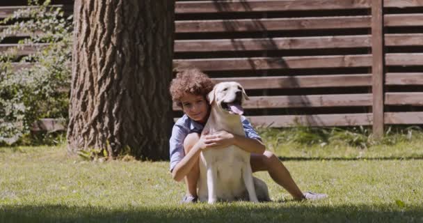 快乐的少年抱着他的狗，享受夏天的户外散步，坐在绿草上，对着相机微笑 — 图库视频影像