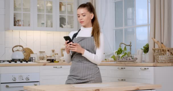 Przepisy online. młoda kaukaska gospodyni domowa noszenie fartuch stojąc w kuchni i networking na smartfon — Wideo stockowe