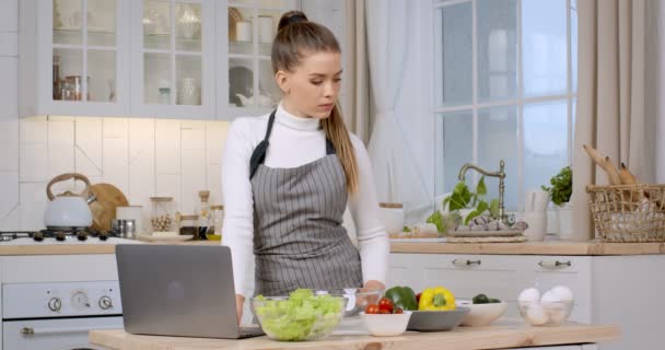 Çevrimiçi aşçılık kursları. Genç beyaz kadın dizüstü bilgisayara bakıyor ve mutfaktaki tüm ürünlerin tariflerini kontrol ediyor. — Stok video