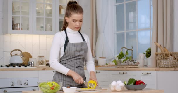Önlük giymiş genç bir bayan taze dolmalık biber kesip kaseye ekliyor, mutfakta sebze salatası pişiriyor. — Stok video