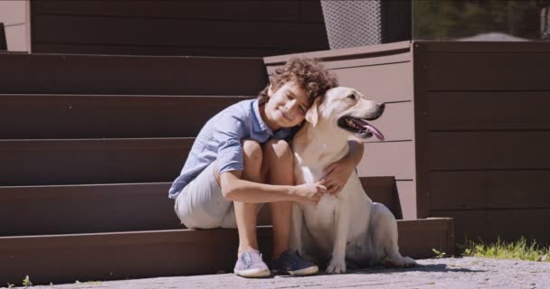 小男孩抱着他可爱的拉布拉多犬，坐在屋外的楼梯上，爱抚着他的朋友 — 图库视频影像