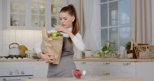 Эко шоппинг. Девушка в фартуке распаковывает покупки из бумажного пакета, кладет органические овощи в миску на кухне — стоковое видео