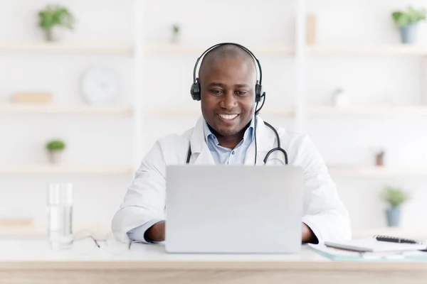 Consulta online durante a quarentena. Médico negro alegre usando fones de ouvido e conversando com o paciente via laptop — Fotografia de Stock