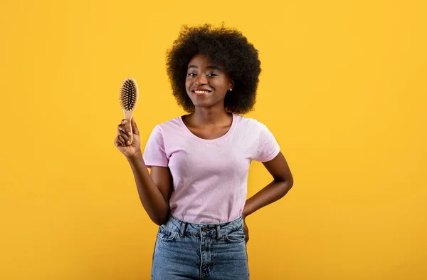 幸せなアフリカ系アメリカ人女性木製の毛ブラシを保持し、黄色のスタジオの背景にカメラに笑みを浮かべて — ストック写真