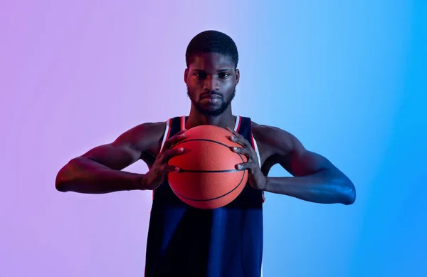Retrato de determinado jovem basquetebolista preto segurando bola e olhando para a câmera em luz de néon — Fotografia de Stock