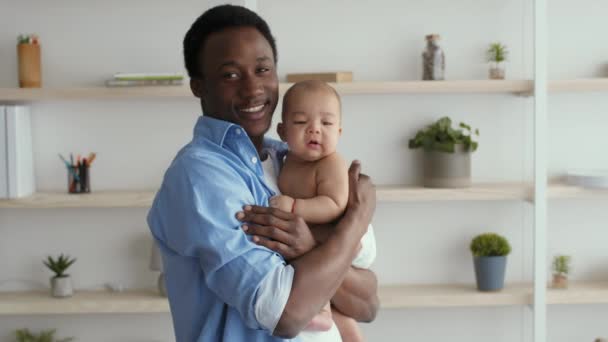 Fröhlich afrikanisch-amerikanischer Papa hält und wiegt sein entzückendes Neugeborenes, lächelt in die Kamera im Wohnzimmer — Stockvideo