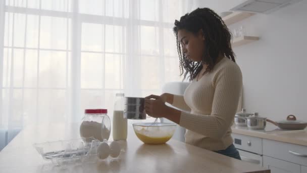 Young concentrated african american woman cooking at kitchen, mixing dough in bowl and adding some flour with sifter — Stock Video