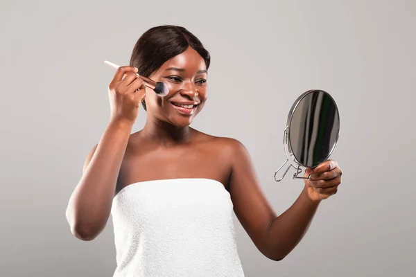Glückliche junge schwarze Frau hält Schminkpinsel und blickt in den Spiegel — Stockfoto