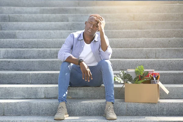 Krise und Arbeitslosigkeit. Gestresster schwarzer Typ mit persönlichen Sachen sitzt nach dem Verlust seines Arbeitsplatzes im Freien auf der Treppe — Stockfoto