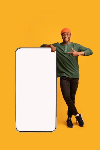 Chico africano alegre inclinándose y apuntando a un teléfono inteligente grande con pantalla blanca —  Fotos de Stock