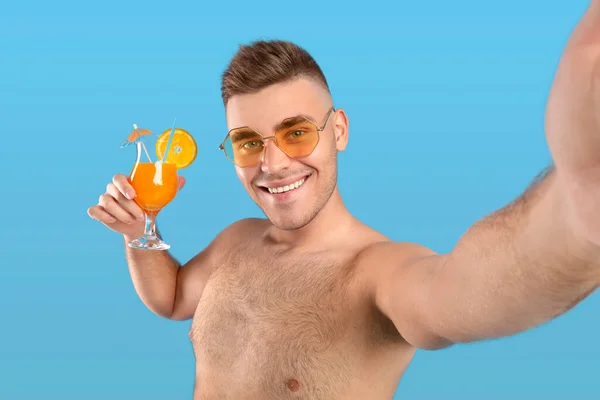 Lächelnder Millennial-Typ mit Sonnenbrille hält kühlen orangefarbenen Cocktail in der Hand und macht Selfie auf blauem Studiohintergrund — Stockfoto