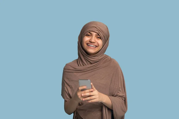 Potret gadis remaja yang bahagia dalam jilbab Islam menggunakan smartphone atas latar belakang studio biru — Stok Foto