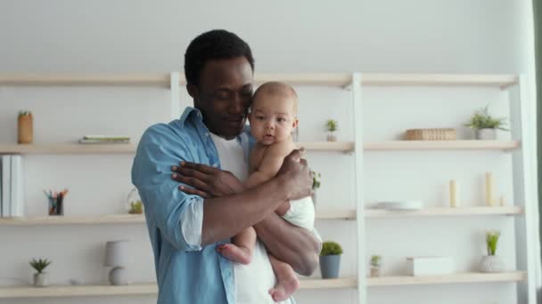 Happy african american dad carrying his newborn baby wearing diaper and talking to him, standing at living room — Video